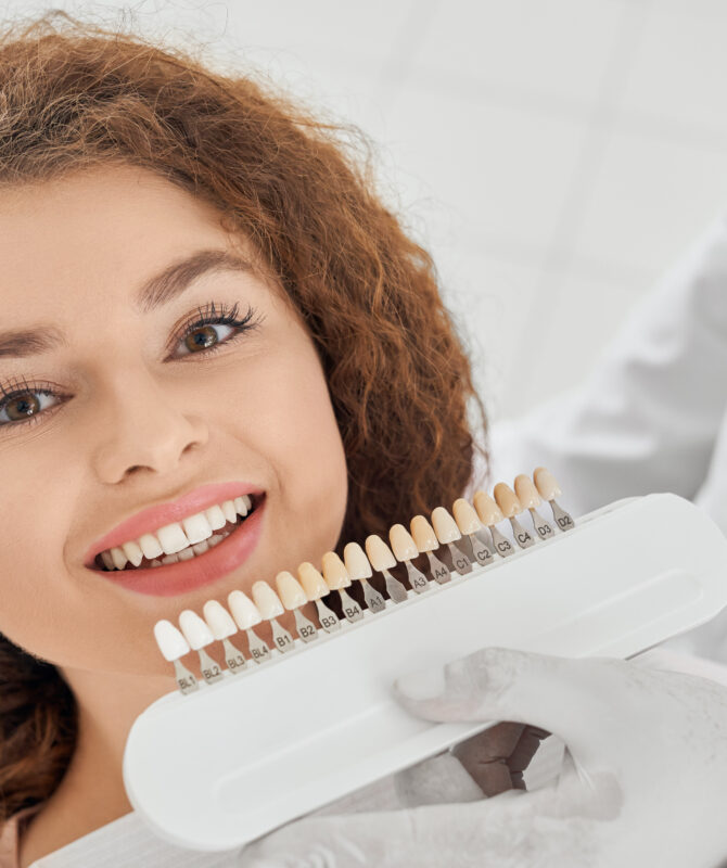 Closeup of beautiful young woman lying on dental chair, looking at camera and smiling while male dentist keeping in hands teeth color range. Girl doing whitening procedure in dental office.