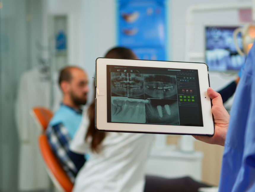 Close up dentist nurse holding tablet with digital radiography, while doctor is working with patient in background examining teeth problem sitting on stomatological chair in dental clinic.